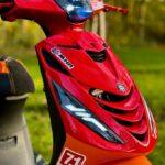 Close-up van de voorkant van een rood-oranje scooter met LED-dagrijlichten en stickers, buiten in een groene omgeving.