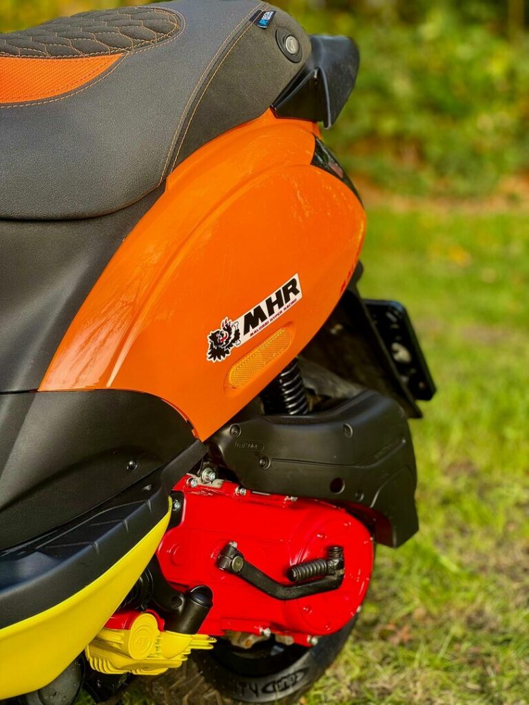 Close-up van een kleurrijke scooter met oranje kuip, rood motorblok en MHR-sticker op het gras.