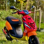 Rood-oranje scooter met sportieve stickers geparkeerd op gras in een bosrijke omgeving.