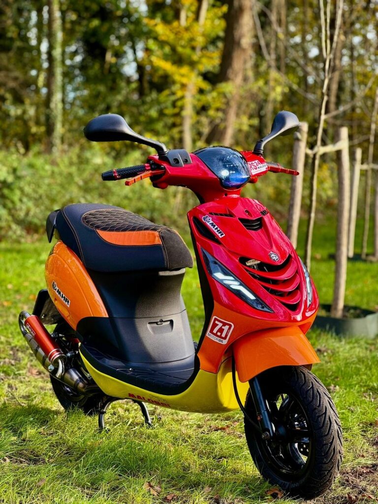 Rood-oranje scooter met sportieve stickers geparkeerd op gras in een bosrijke omgeving.