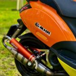 Close-up van de achterzijde van een oranje scooter met Yasuni sportuitlaat en zwart zadel, geparkeerd op gras.