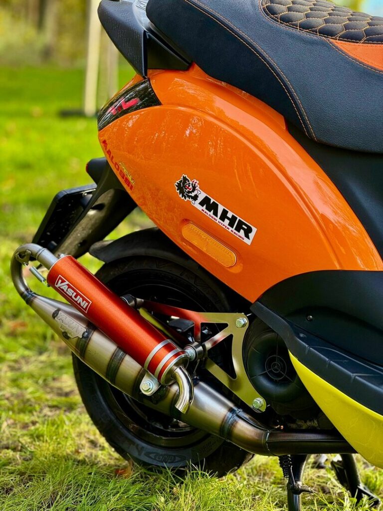 Close-up van de achterzijde van een oranje scooter met Yasuni sportuitlaat en zwart zadel, geparkeerd op gras.