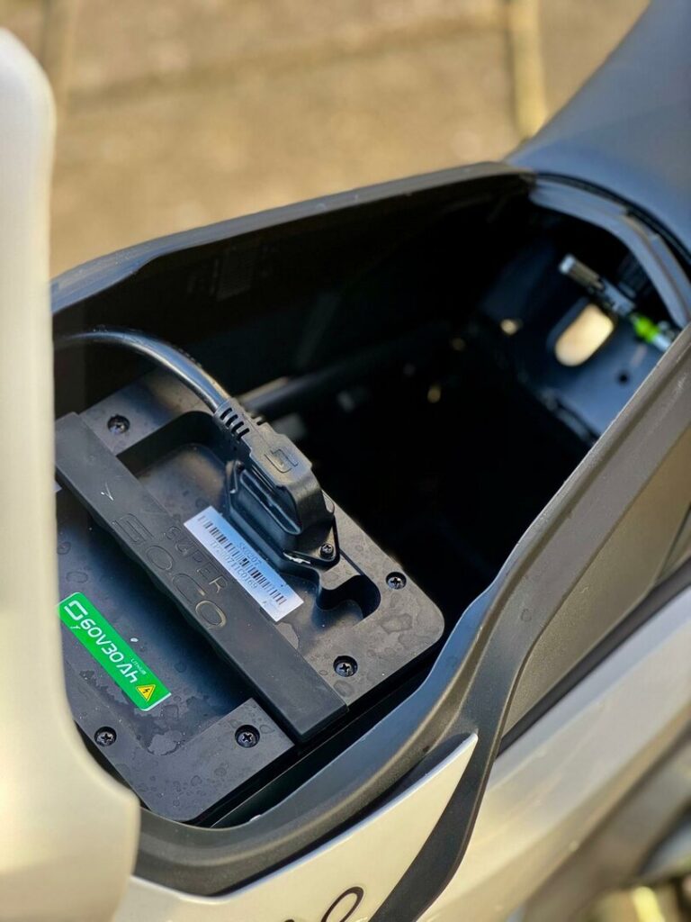 Close-up van de Super Soco elektrische scooteraccu met aangesloten laadkabel in het zadelvak.