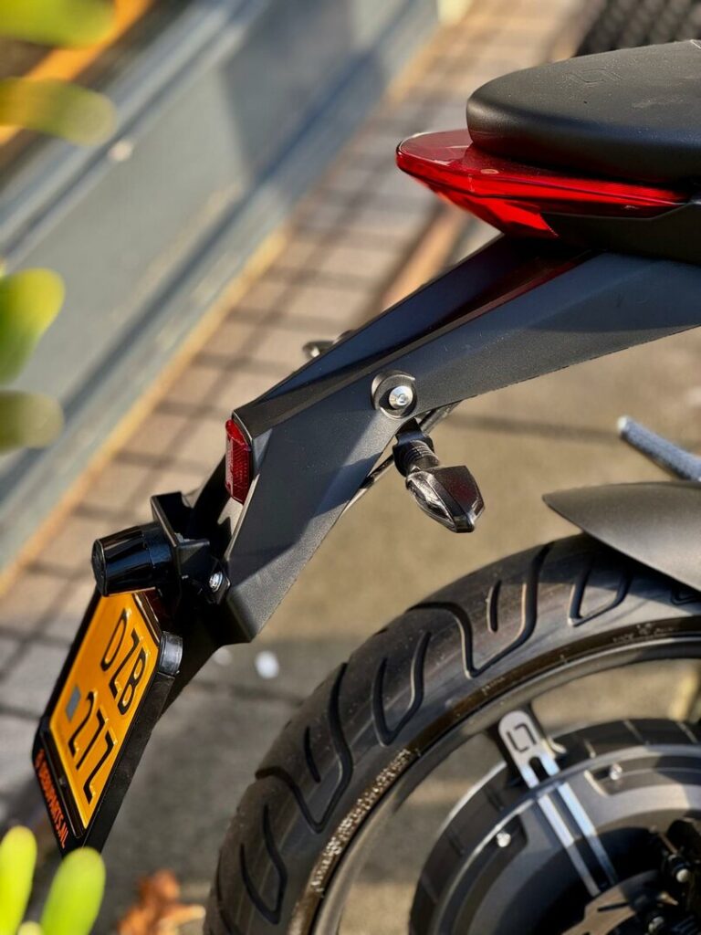 Achterzijde van een motorfiets met rode achterlicht, knipperlicht en gele kentekenplaat.