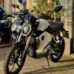 Elektrische motorfiets van Super Soco geparkeerd voor een showroom, in de zon naast andere scooters.