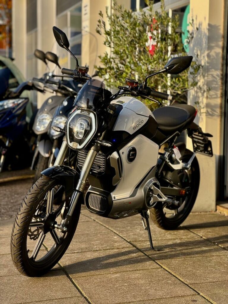 Zilverkleurige Super Soco elektrische motor op standaard, voor een showroom met scooters op de achtergrond.