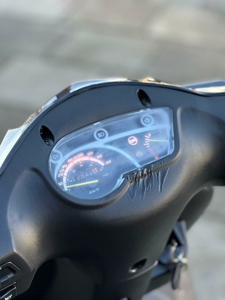 Close-up van een scooterdashboard met snelheidsmeter en gescheurde, vervormde kunststof kap.