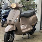Beige retro scooter met brandende koplamp, geparkeerd voor een winkel met rek achterop.