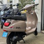 Beige agm VX50 bromscooter met bagagedrager, standpoot en blauwe NL-kentekenplaat, geparkeerd bij winkel