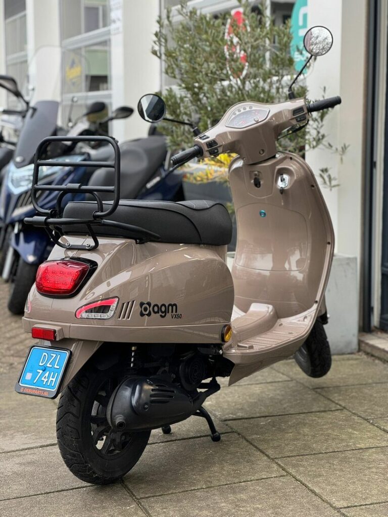Beige agm VX50 bromscooter met bagagedrager, standpoot en blauwe NL-kentekenplaat, geparkeerd bij winkel