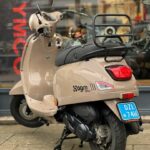 Beige AGM VX50 scooter met bagagerek, middenstandaard en NL kenteken, geparkeerd voor winkelraam.