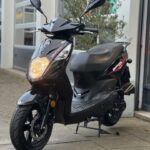 Zwarte SYM Orbit II scooter met brandend voorlicht, geparkeerd op stoep bij garage.