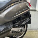 Close-up van de achterzijde van een grijze scooter met zadel, uitlaat en chromen bagagedrager.