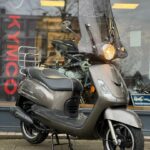 Grijze retro-scooter met hoog windscherm en brandende koplamp voor een winkel met KYMCO-logo.