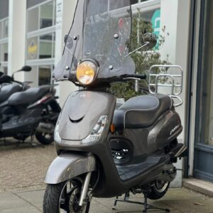 Grijze scooter met hoog windscherm en brandende koplamp, geparkeerd voor een winkel.