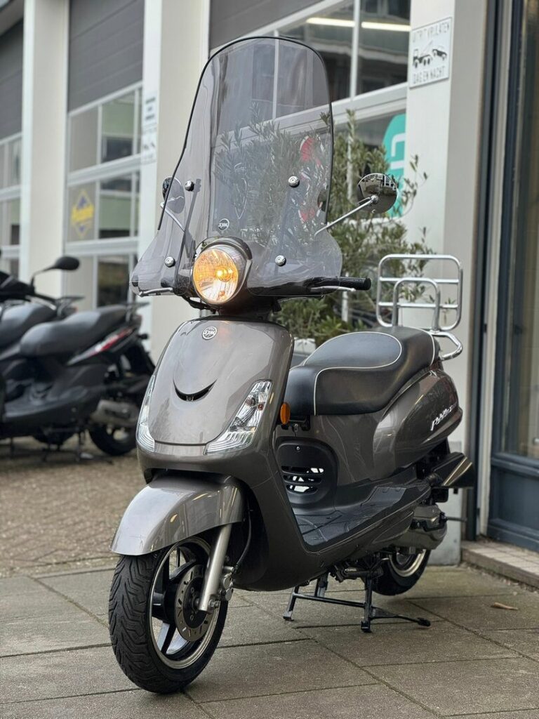 Grijze scooter met hoog windscherm en brandende koplamp, geparkeerd voor een winkel.