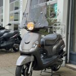 Grijze scooter met hoog windscherm en brandende koplamp, geparkeerd voor een werkplaats.