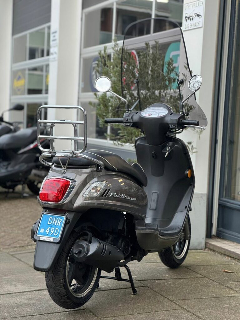 Grijze scooter met windscherm en chromen bagagedrager, geparkeerd op de stoep voor een winkel.