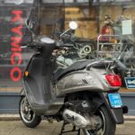 Grijze Kymco-scooter met hoog windscherm en bagagedrager, geparkeerd voor een showroom.
