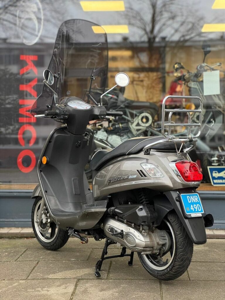 Grijze Kymco-scooter met hoog windscherm en bagagedrager, geparkeerd voor een showroom.