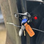 Close-up van een scooterslot met sleutelbos en leren 'Vespa' sleutelhanger aan een zwarte scooter.