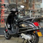 Zwarte Vespa scooter met chromen bagagerek en windscherm, geparkeerd voor een showroom.