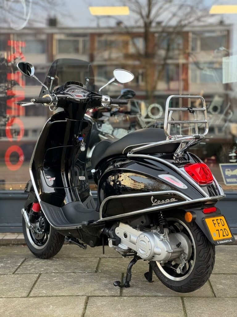 Zwarte Vespa scooter met chromen bagagerek en windscherm, geparkeerd voor een showroom.