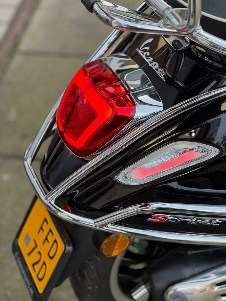 Close-up van de achterkant van een zwarte Vespa Sprint met rood achterlicht, chroomrek en gele kentekenplaat.