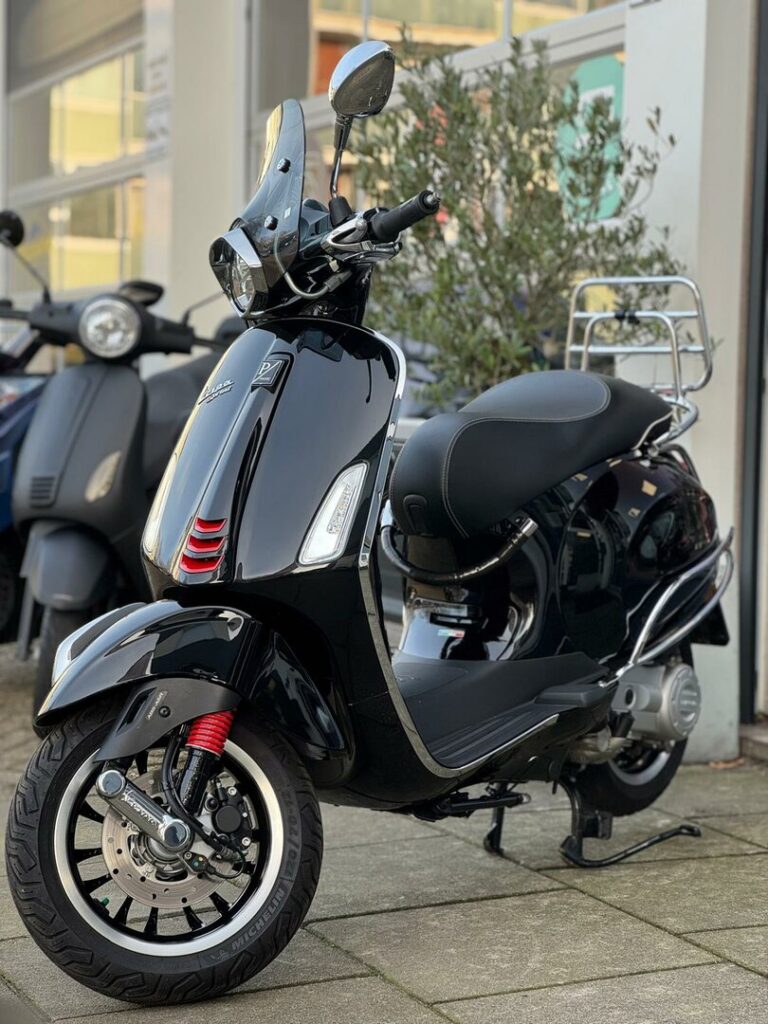 Glanzend zwarte scooter met ruitje en rood geveerde voorvork, geparkeerd bij een winkel.
