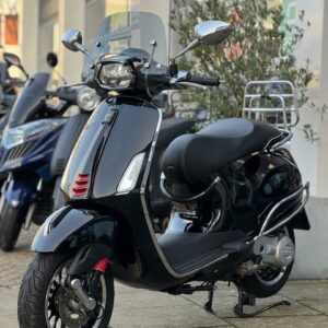 Zwarte Vespa-scooter met windscherm en chromen details, geparkeerd voor een winkel.