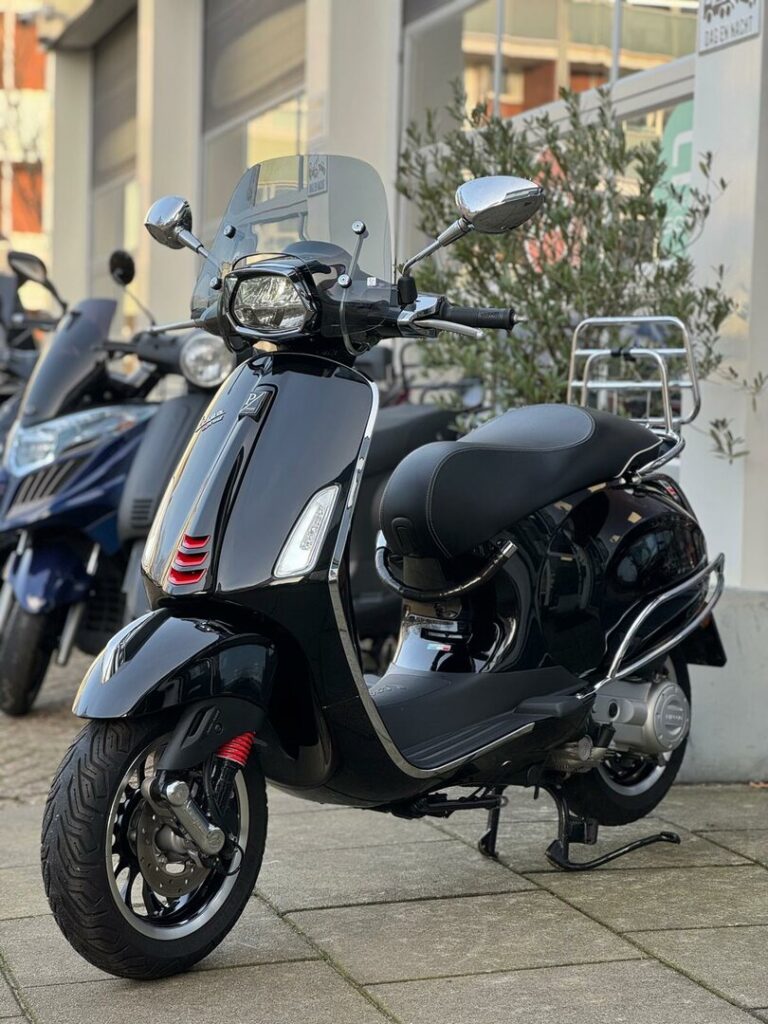 Zwarte Vespa-scooter met windscherm en chromen details, geparkeerd voor een winkel.