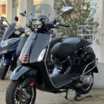 Zwarte Vespa-scooter met windscherm en bagagedrager, op standaard geparkeerd bij showroom.