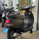 Zwarte scooter met bagagedrager, op standaard geparkeerd voor een scooterdealer.