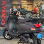 Mat zwarte scooter op middenbok met bagagedrager, voor winkelpui met KYMCO-logo.