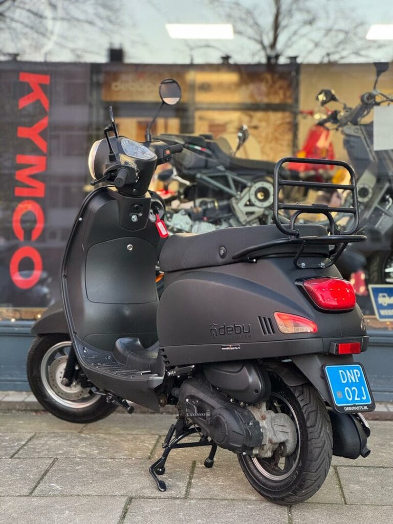 Mat zwarte scooter op middenbok met bagagedrager, voor winkelpui met KYMCO-logo.