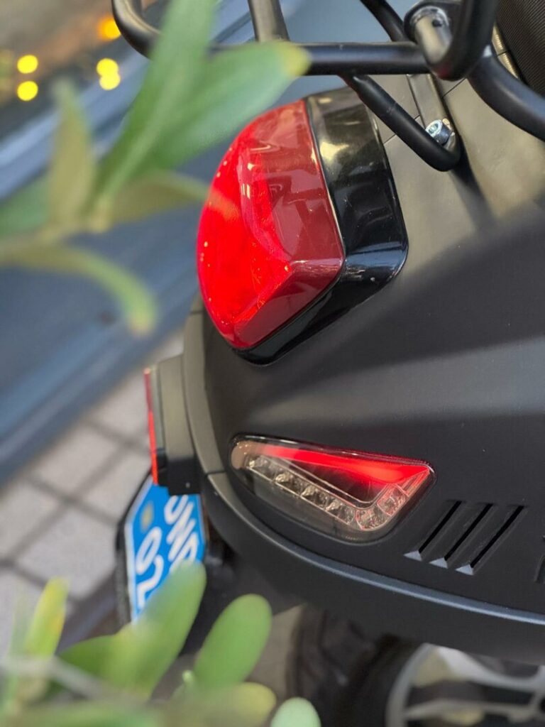 Close-up van de achterzijde van een scooter met brandend achterlicht en knipperlicht, blauwe kentekenplaat.