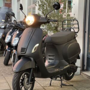 Matzwarte scooter met brandende koplamp op middenstandaard, geparkeerd voor een winkel met andere scooters.