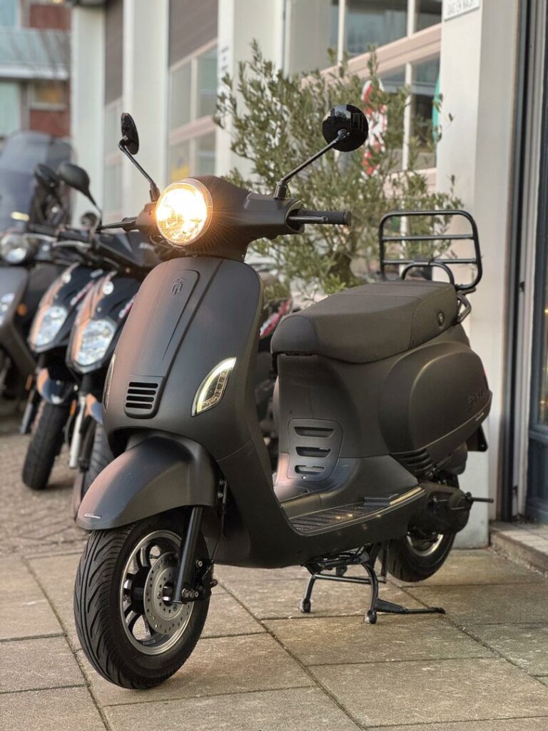 Matzwarte scooter met brandende koplamp op middenstandaard, geparkeerd voor een winkel met andere scooters.