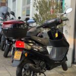 Zwarte scooter met rode topkoffer, geparkeerd op de stoep bij een werkplaats.