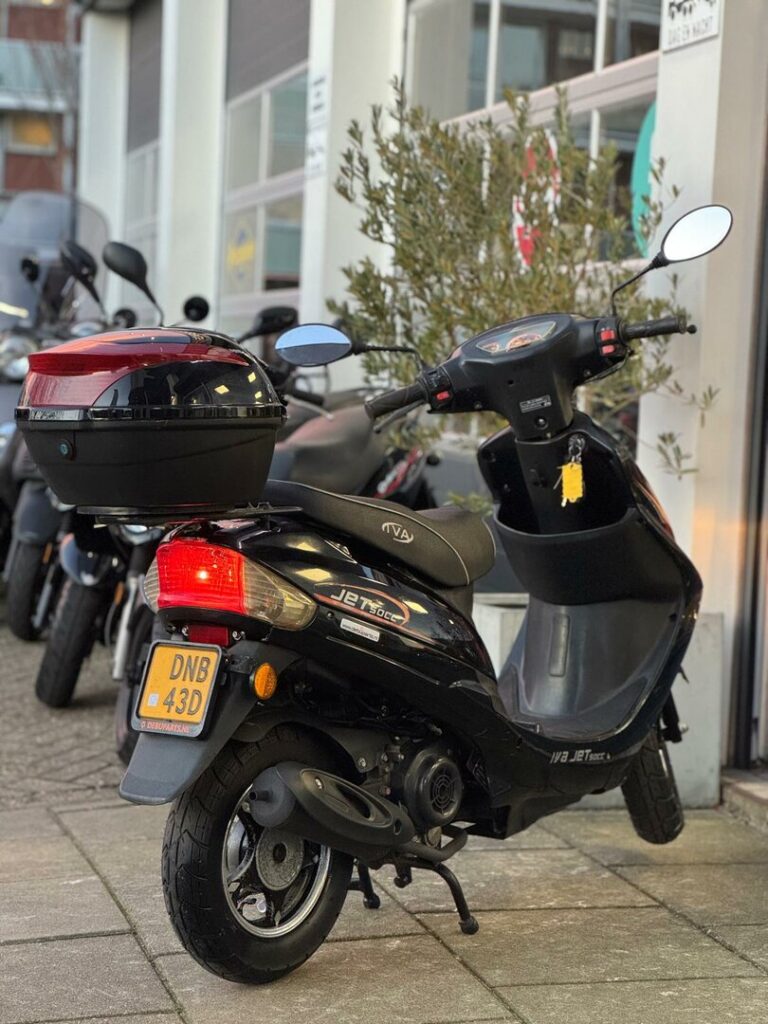 Zwarte scooter met rode topkoffer, geparkeerd op de stoep bij een werkplaats.