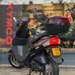 Zwarte scooter met topkoffer geparkeerd voor een motorzaak met KYMCO-logo.