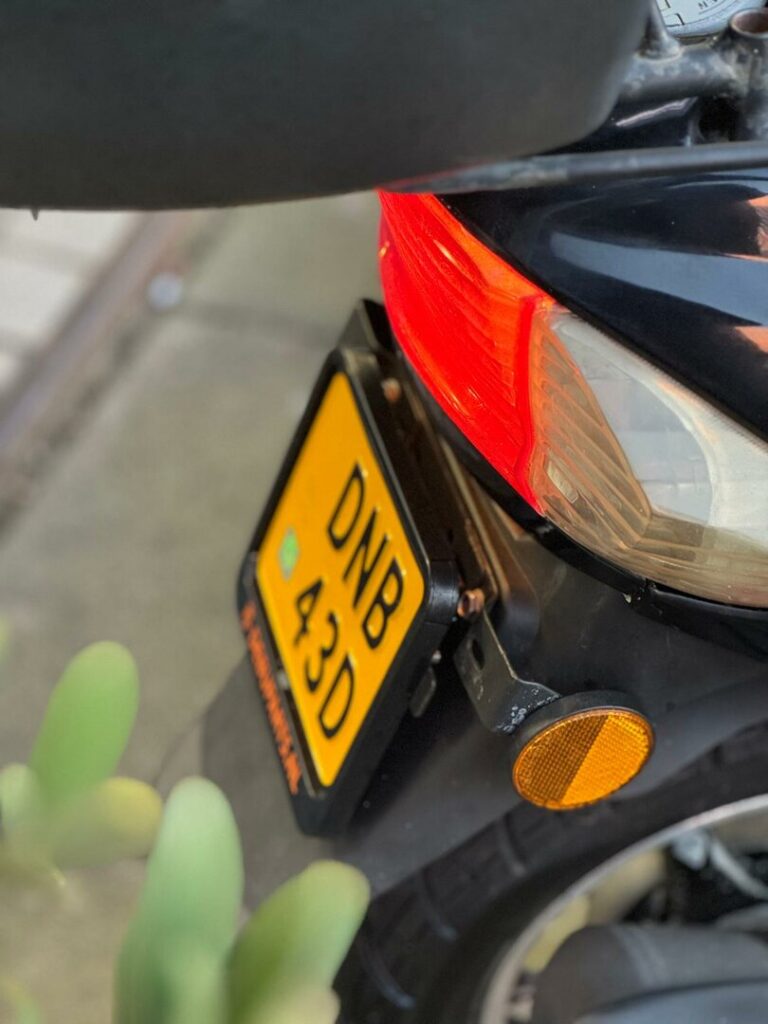Close-up van de achterkant van een motorfiets met geel kenteken, achterlicht en oranje reflector.