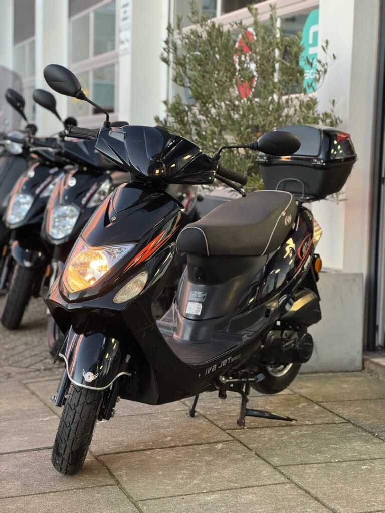 Zwarte scooter met topkoffer voor een winkel, koplamp aan; andere scooters op de achtergrond.