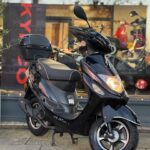 Zwarte scooter met topkoffer geparkeerd voor een winkelraam, met brandende koplamp.