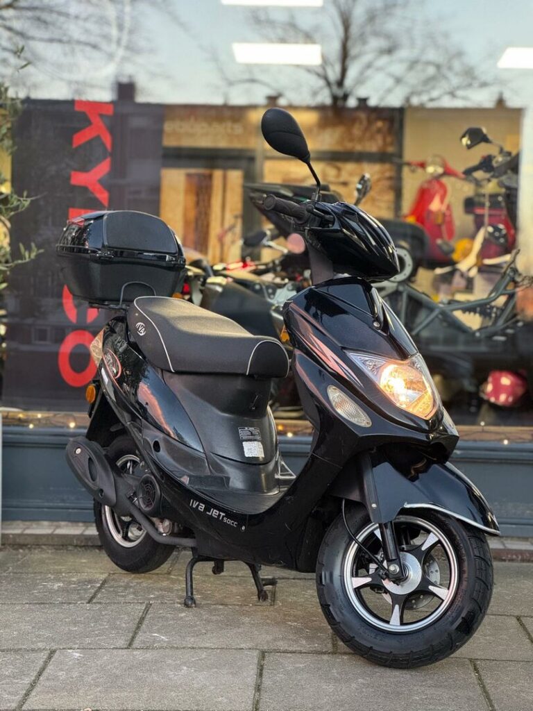 Zwarte scooter met topkoffer geparkeerd voor een winkelraam, met brandende koplamp.