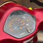 Close-up van de snelheidsmeter, kilometerteller en klokje op een rode scooter, bedekt met regendruppels.