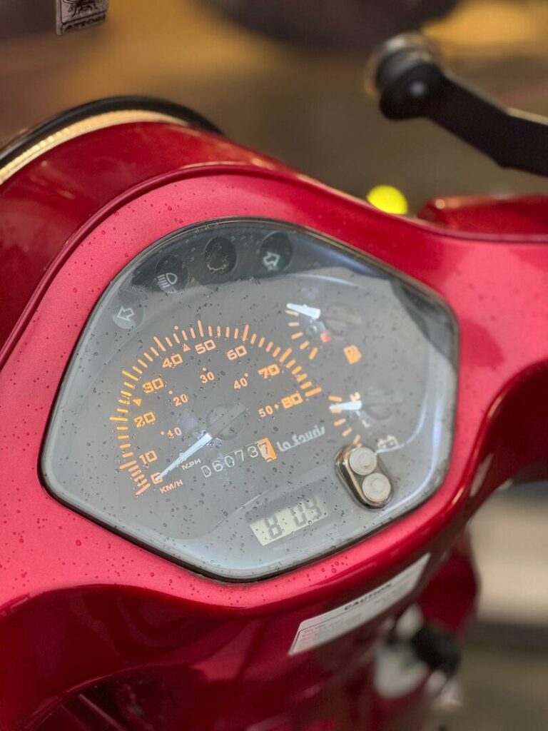 Close-up van de snelheidsmeter, kilometerteller en klokje op een rode scooter, bedekt met regendruppels.