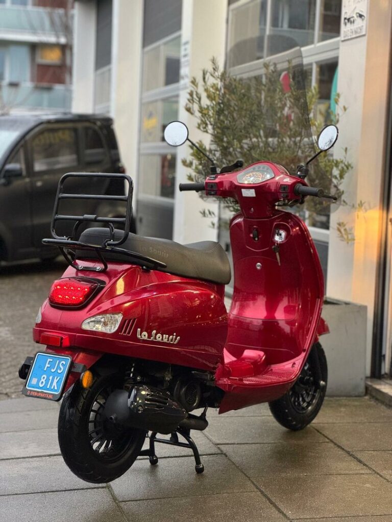 Rode La Souris retro-scooter geparkeerd op nat trottoir voor een garage.