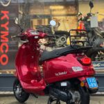 Rode La Souris-scooter met bagagerek voor een motorzaak, geparkeerd op de stoep.