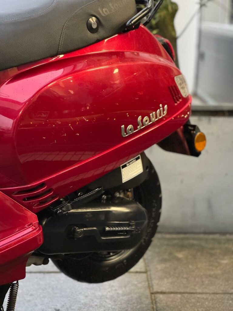 Close-up van de rode La Souris-scooter, met krassen op de zijkap en zicht op zadel en achterwiel.